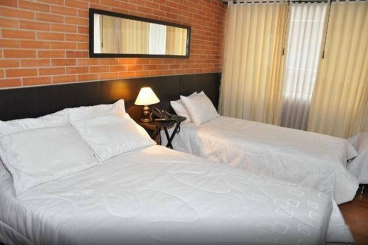 Habitación de hotel con dos camas individuales, una mesita de noche con lámpara, un espejo en la pared de ladrillo y una ventana con cortinas.