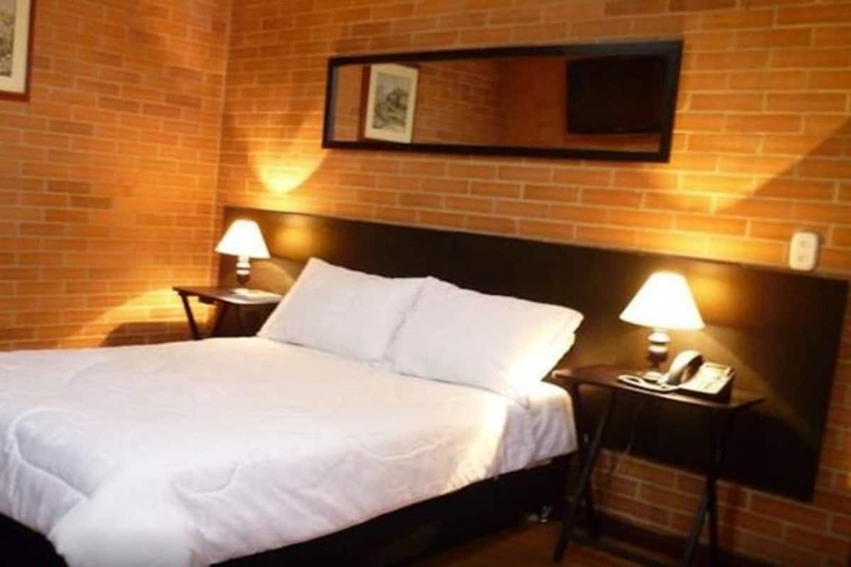 Habitación de hotel con pared de ladrillo, una cama con ropa de cama blanca, dos mesitas de noche con lámparas y cuadros en la pared.