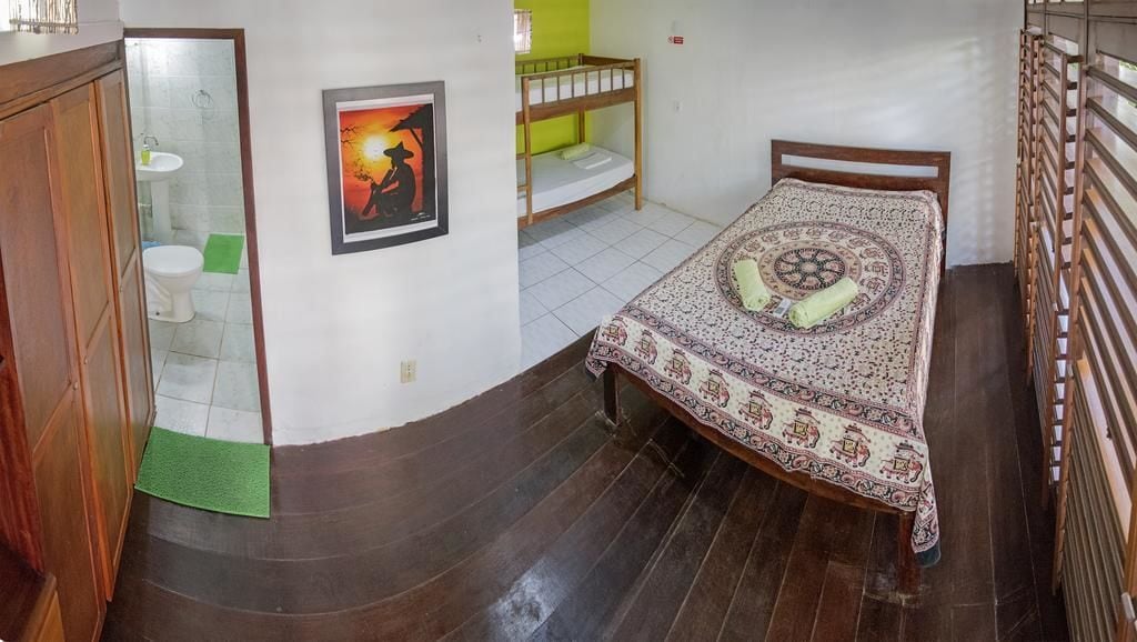 A small, compact room with a single bed, a wooden wardrobe, a green mat, a framed picture, and a bunk bed in the background.