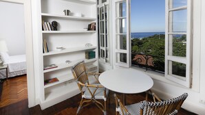 A room with a white interior, a round table, two wicker chairs, a bookshelf, and a large window with a view of the sea. - Rio107 - Apartment Copacabana 2 Bedrooms (Rio de Janeiro)