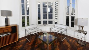 A corner room with a glass-top table, two white armchairs, a wooden cabinet with electronic devices, and large windows offering a view of buildings. - Rio107 - Apartment Copacabana 2 Bedrooms (Rio de Janeiro)