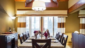 A dining area with a table set for four, wooden chairs, a chandelier, and large windows with curtains.