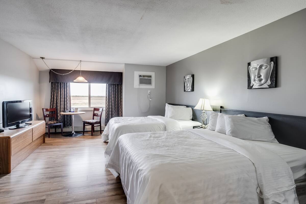 A hotel room with two beds, a desk with a TV, a dining area with chairs, and a window with curtains.