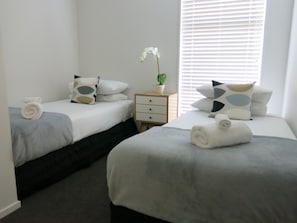 A hotel room with two beds, each with a rolled towel, a potted plant, and patterned pillows. There is a small bedside table with a drawer and a nightstand with a lamp.