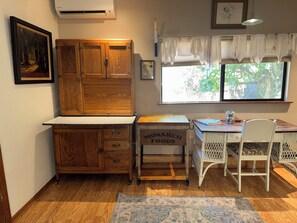 A kitchen with wooden cabinets, a white countertop, a wicker chair, and a window with curtains. - The Vintage Cottage with riverfront access (Grants Pass)