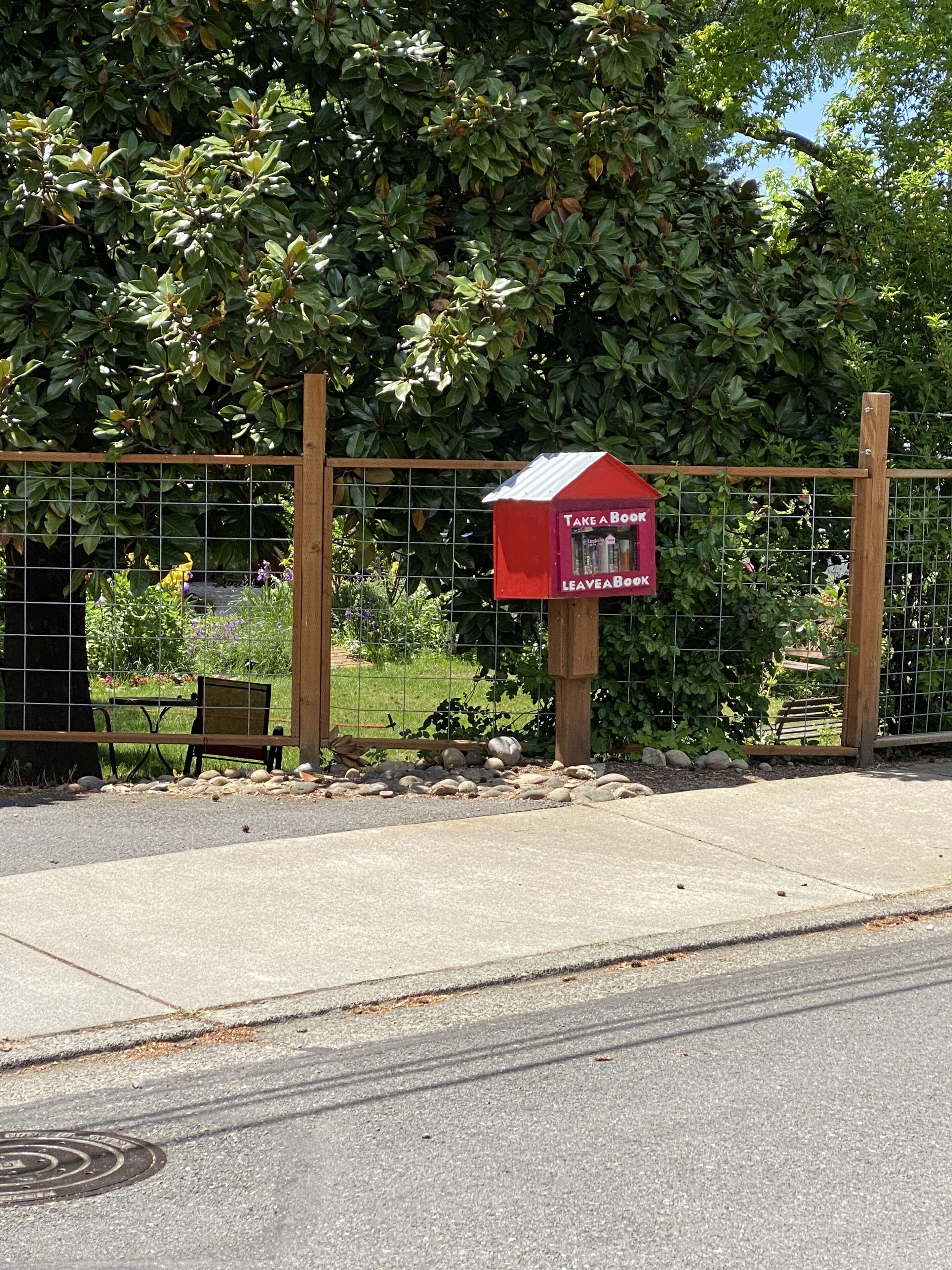 Une boîte aux lettres rouge avec une inscription indiquant « Empruntez un livre, déposez un livre », une clôture en bois et un banc, le tout situé dans un jardin.
