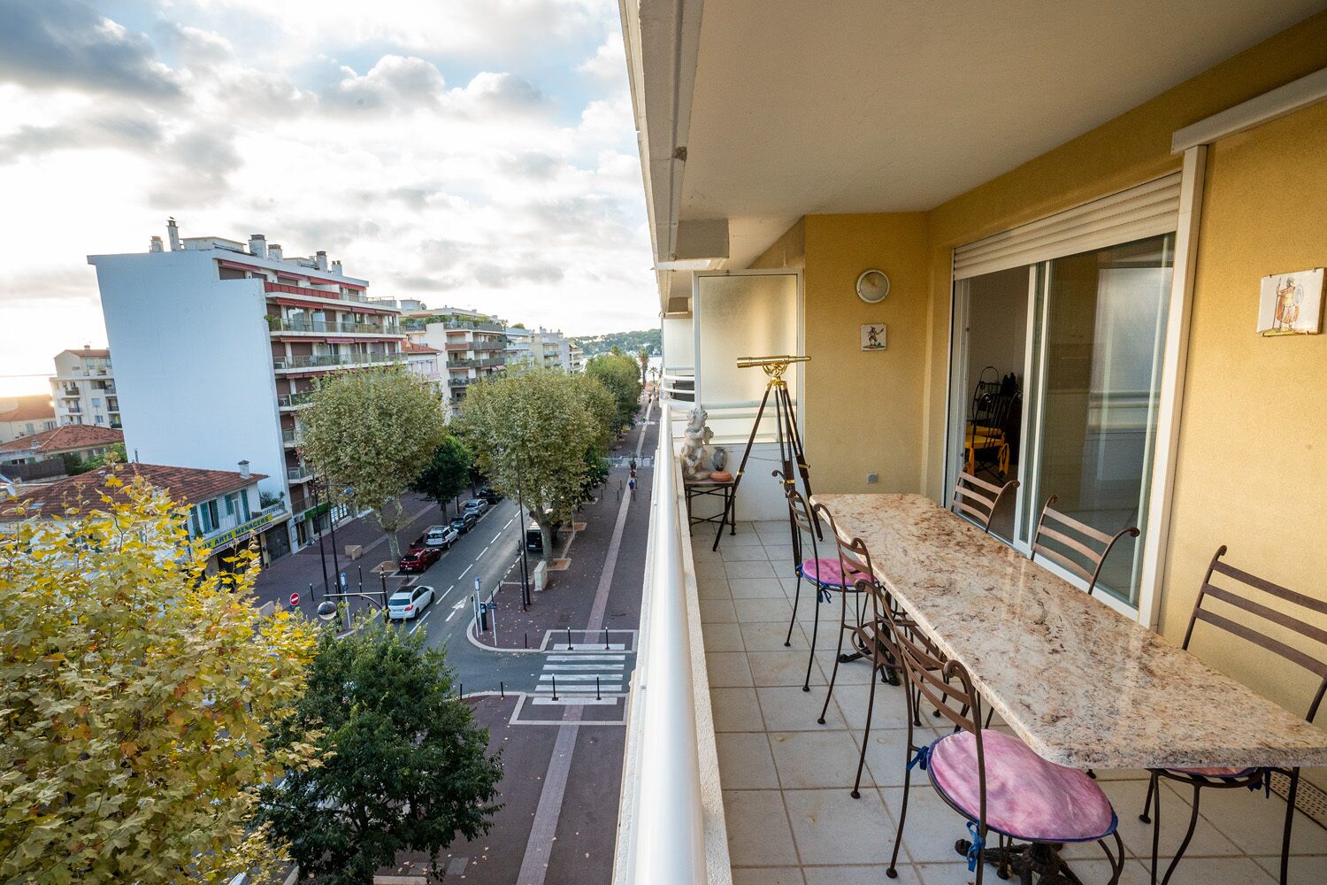 Un balcón con mesa y sillas de mármol, que da a una calle con árboles y edificios.