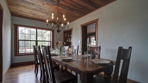 A dining room with a wooden table set for four, surrounded by chairs. A chandelier hangs from the ceiling, and there are windows providing natural light.