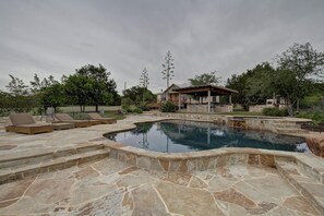 A stone pool with steps leading into it, surrounded by lounge chairs and a wooden deck. - Beautiful 5 Acre Hill Country Retreat with Pool, Hot Tub and Outdoor Kitchen!!!  (Buda)