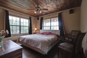 Un dormitorio con techo de madera, una cama con cubrecama blanco, dos sillas, una mesita de noche con lámpara y una ventana con cortinas.