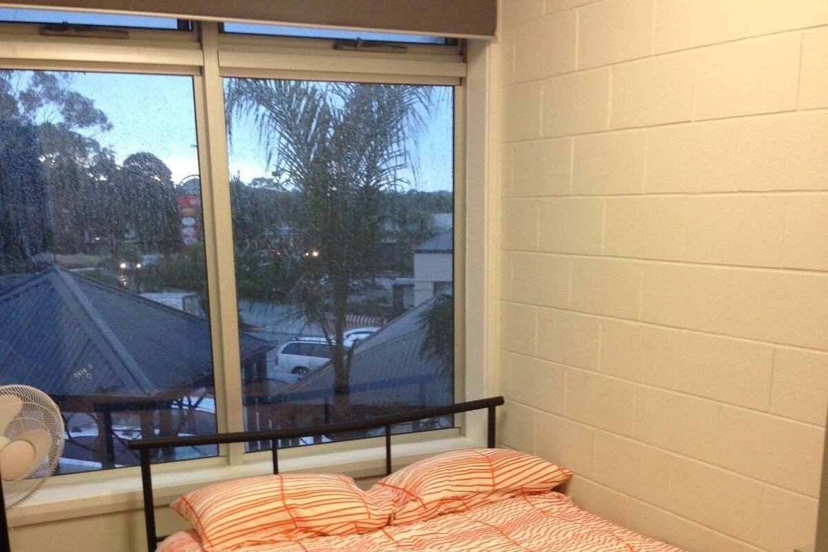 A single bed with a striped blanket in a room with a window overlooking a parking area and trees.