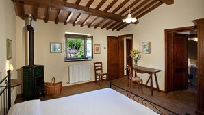 Un dormitorio con cama, un aparador de madera, una ventana con vista a la vegetación, una mesa de comedor con sillas y un baño visible a través de una puerta abierta.