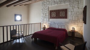 A bedroom with a stone wall, a bed with a red bedspread, a chair, a nightstand, and a small table with a lamp. - AGRITURISMO I5SENSI (Vc) Studio of 45 square meters, with swimming pool. (Modica)