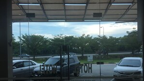A view through a glass door of a parking area with cars parked, a clear sky, and trees in the background. - Ayer Hitam Hotel (Ayer Hitam)