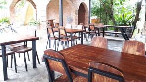 Un área de comedor al aire libre con arcos de ladrillo, mesas y sillas de madera, y plantas en macetas.