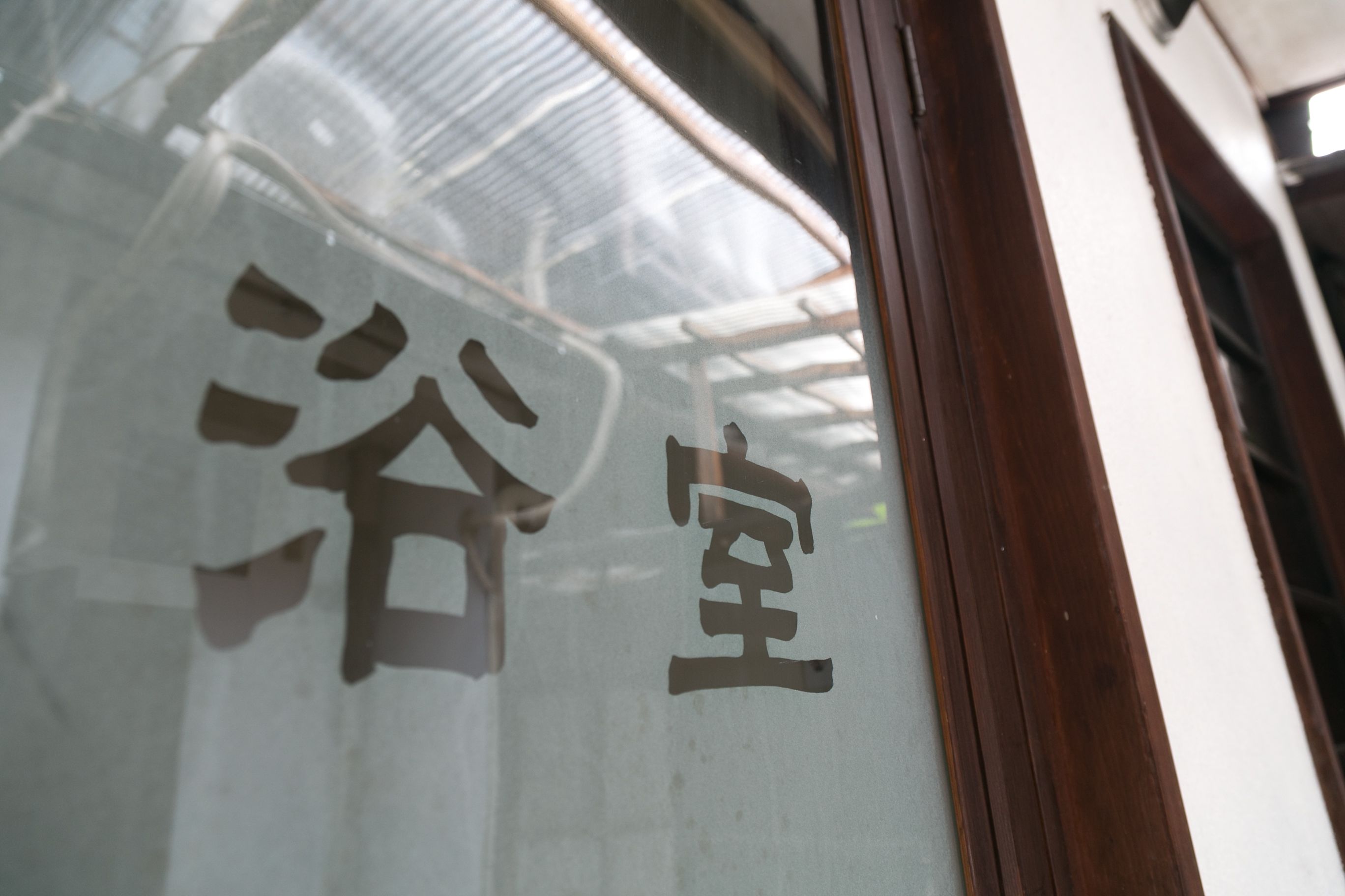 Una ventana de vidrio con el reflejo del interior de un edificio, incluyendo un marco de puerta de madera y una pared blanca.