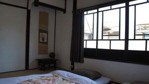 A traditional Japanese-style room with tatami flooring, a low bed with a floral bedspread, a small table with a decorative item, and a large window with a view of the outside. - Guesthouse OKI's Inn - Hostel (Kyoto)