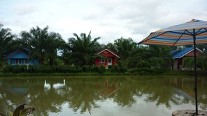 A calm lake with two small houses, one red and one blue, surrounded by lush greenery. - Maneemudjalin Resorts Farm Stay (Sam Roi Yot)