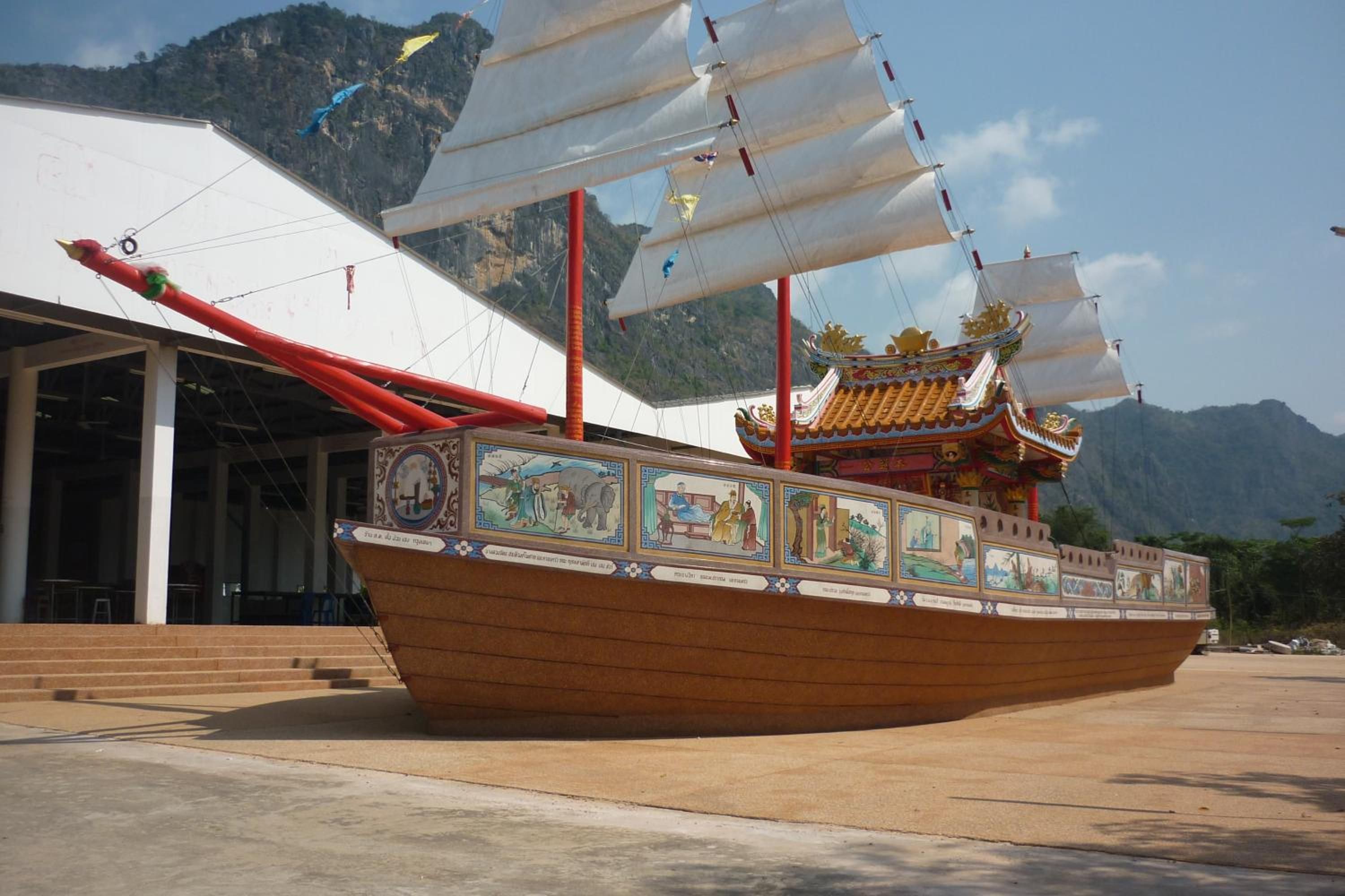 A traditional boat with a decorative roof and painted panels, docked in a concrete area with a mountain backdrop.