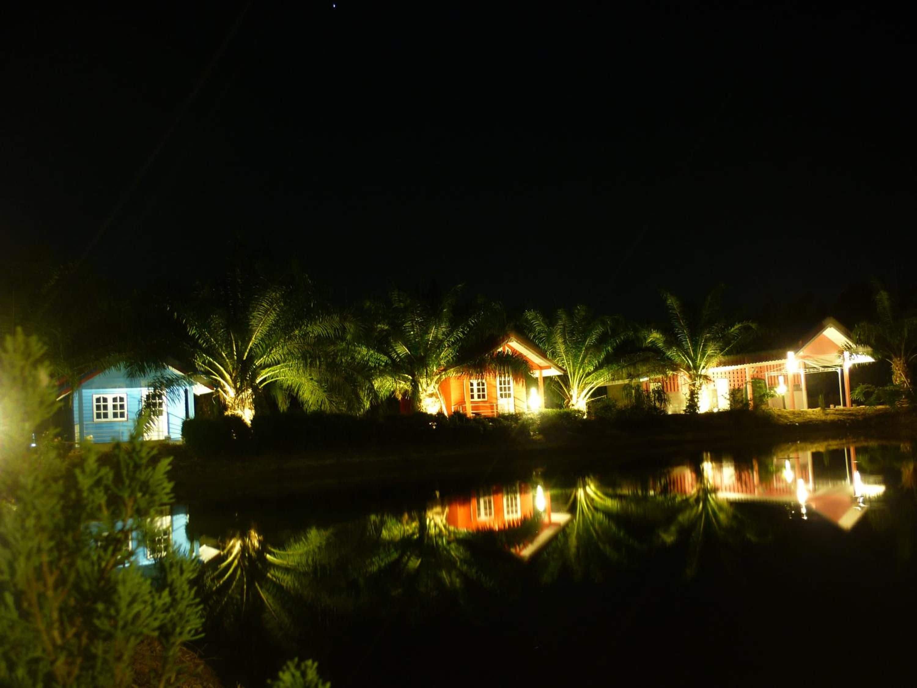A nighttime view of a row of houses with a calm water body in the foreground reflecting the lights.