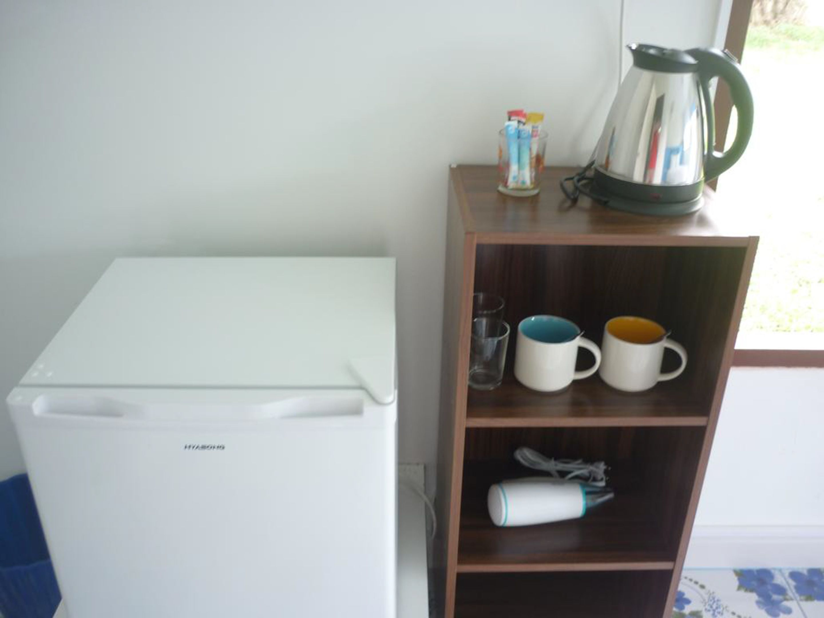 Een kleine, witte koelkast met vriesvak, een houten plankje met drie mokken, een waterkoker en een bak met bestek.
