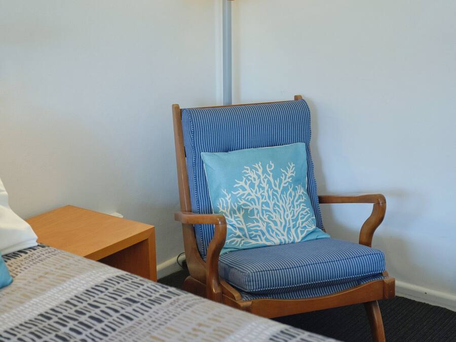 Ett hotellrum med en trästol med en blå kudde, ett sängbord och en lampa med en träbas.