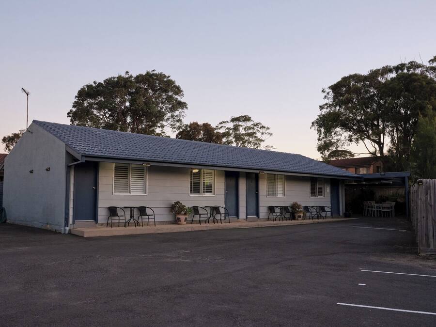 Un motel de una sola planta con techo de tejas, un área de estar al aire libre techada y un estacionamiento cercado.