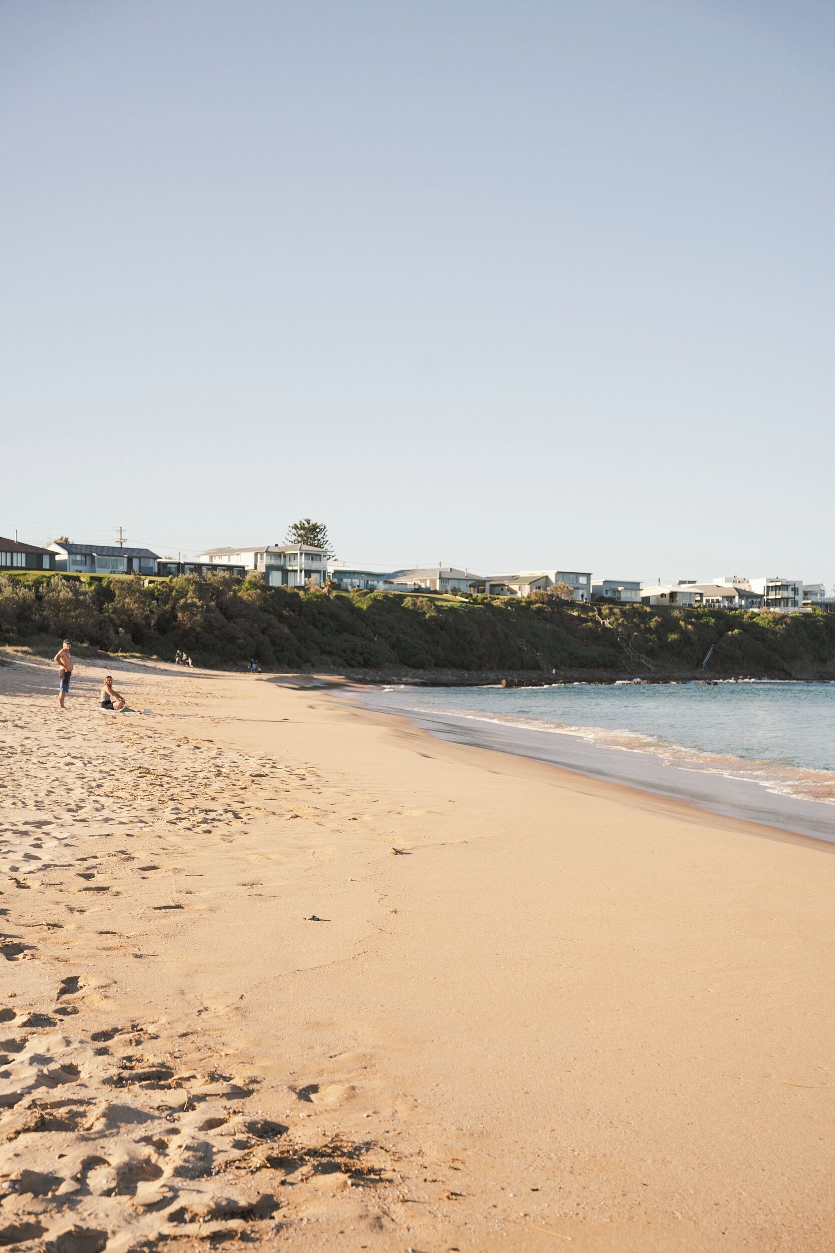 Una playa arenosa con huellas, algunas personas y casas al fondo.