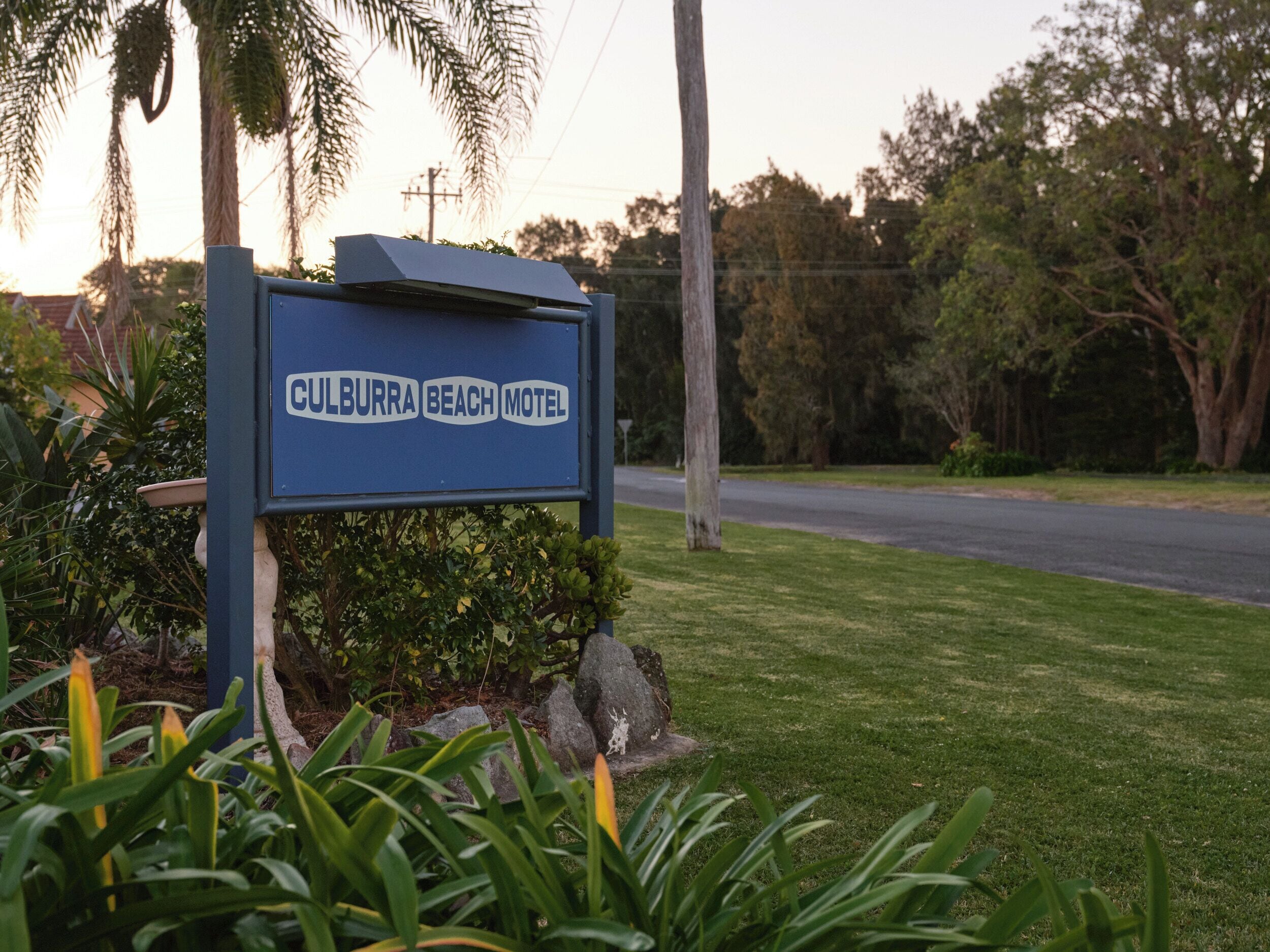 A sign for the Culburra Beach Motel with a solar panel, surrounded by greenery and a road in the background.