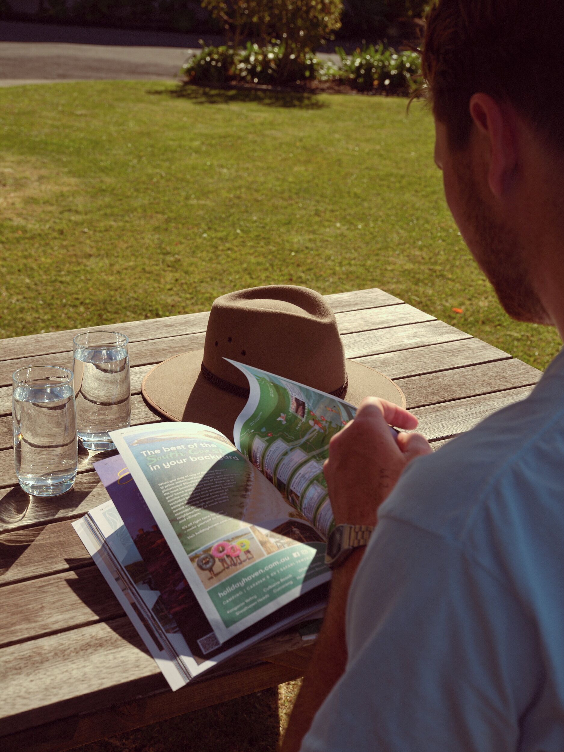 En person som läser en tidning vid ett träbord, och som bär hatt och glasögon.