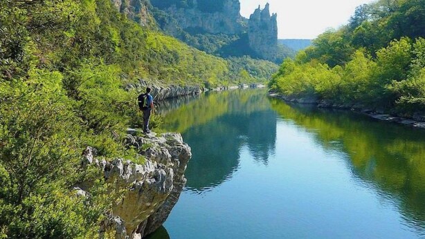 Une personne se tient sur un promontoire rocheux, surplombant une rivière paisible, avec une végétation luxuriante et une montagne en arrière-plan.