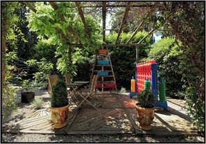 A wooden deck with a ladder leading to a colorful play structure, surrounded by potted plants and greenery. - "Cabanon" - sleeps 2 self catering studio gite 10mins from Avignon! (Rognonas)