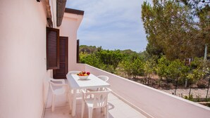 Varanda com mesa e cadeiras brancas, uma tigela de fruta e vista para árvores e uma cerca.