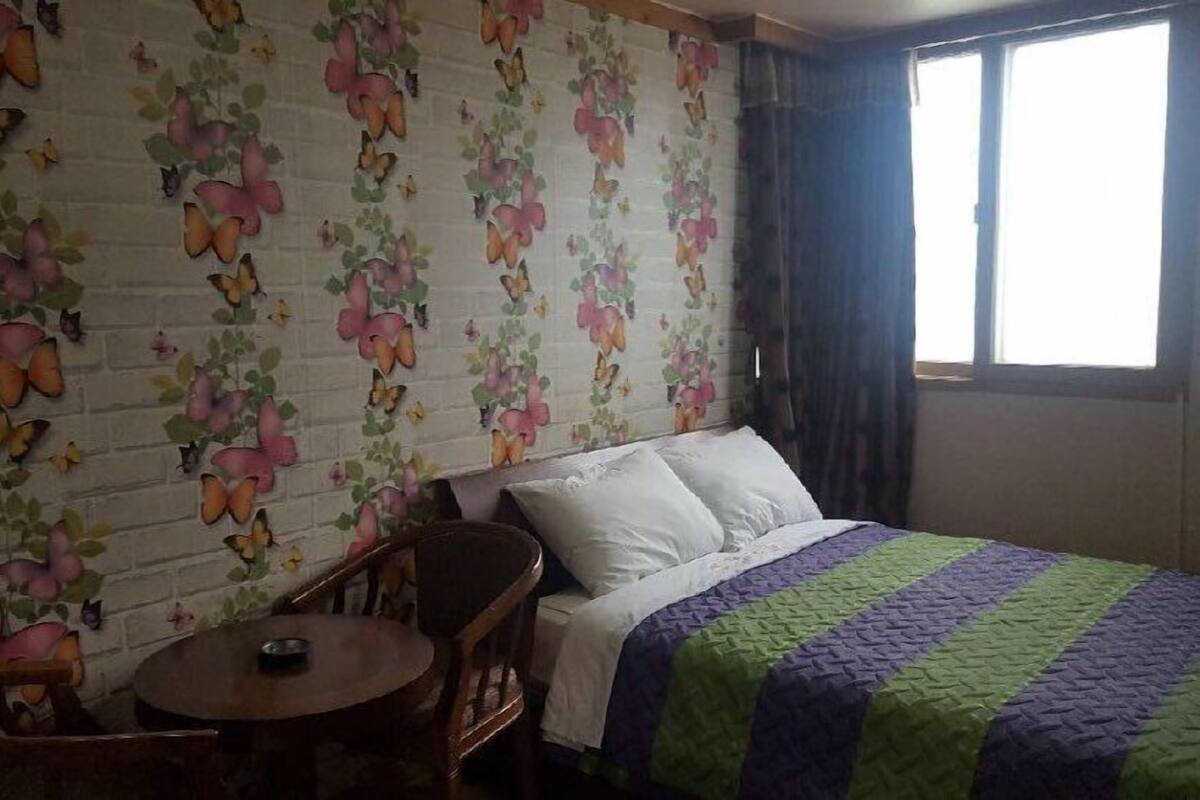 Ein Schlafzimmer mit einer Wand im Backsteinoptik, einem Bett mit gestreifter Decke, einem Holzstuhl und einem kleinen runden Tisch.