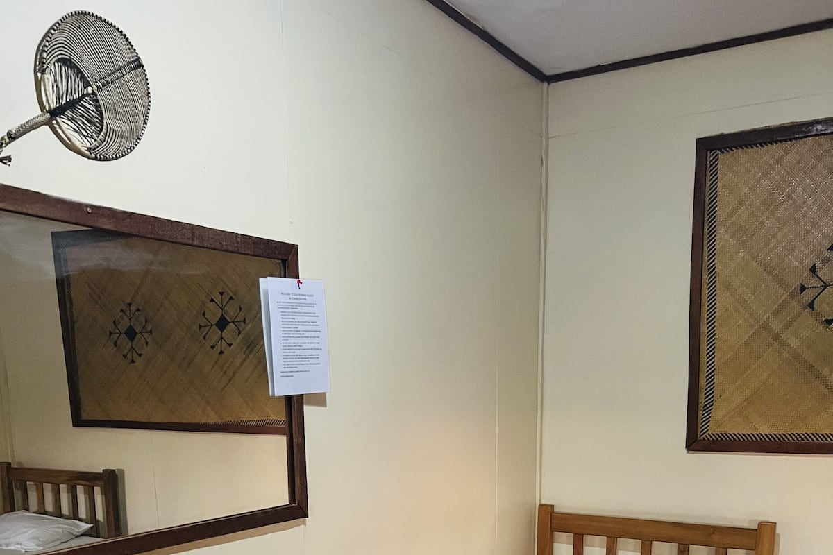 A small, neatly arranged room with a single bed, a wooden desk, a chair, a wall-mounted fan, and a framed picture.