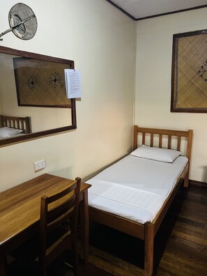 A small, neatly arranged room with a single bed, a wooden desk, a chair, a wall-mounted fan, and a framed picture.