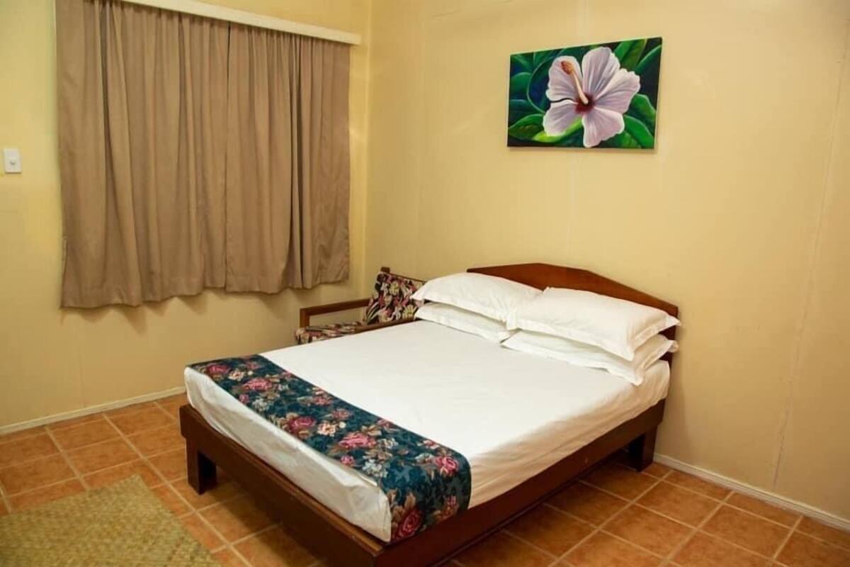 A bedroom with a bed, curtains, a floral painting, and a rug.