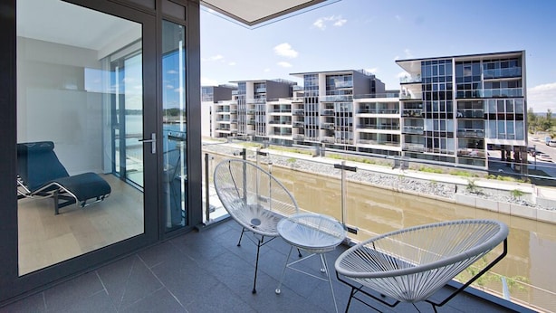 A modern balcony with glass doors, two white wire chairs, and a view of contemporary buildings across a water body. - Accommodate Canberra - Aurora (Kingston)
