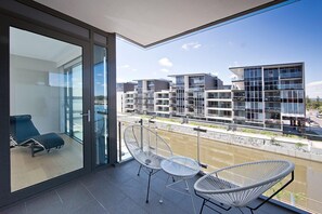 A modern balcony with glass doors, two white wire chairs, and a view of contemporary buildings across a water body.