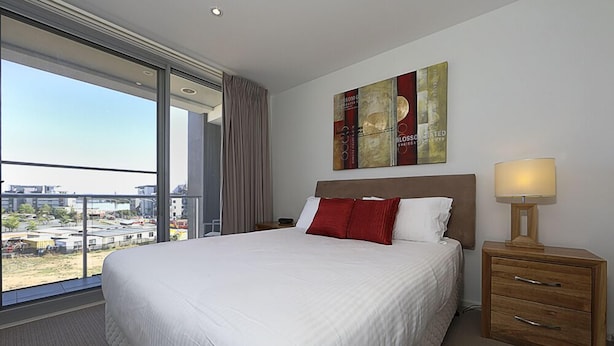 A hotel room with a large bed, a nightstand, a lamp, and a view of the city through a large window. - Accommodate Canberra - Aspire (Kingston)