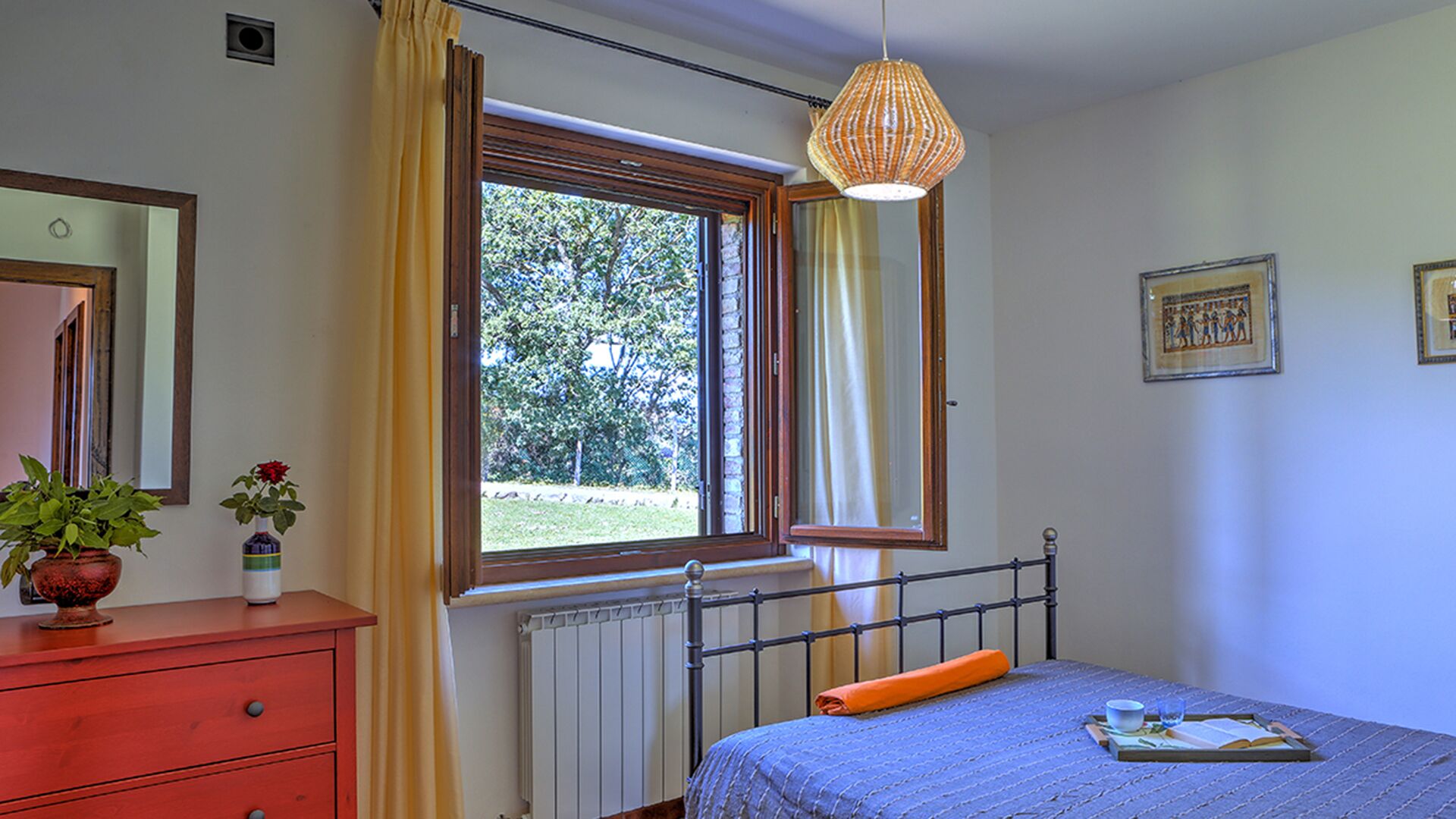 Un dormitorio con ventana, un radiador, una cama con cubierta azul, un aparador rojo y una lámpara colgante.