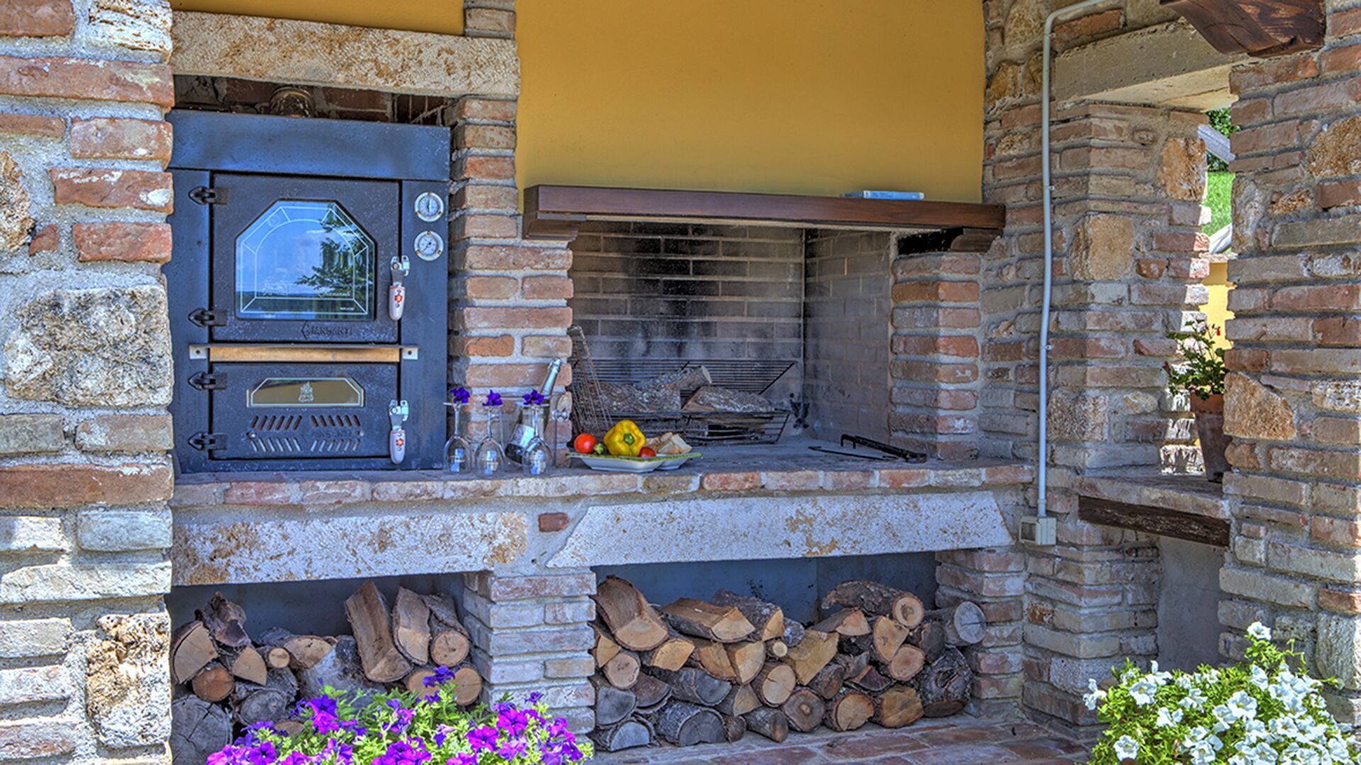 An outdoor brick oven with a wooden shelf above it, a firewood stack below, and a small garden area with flowers.