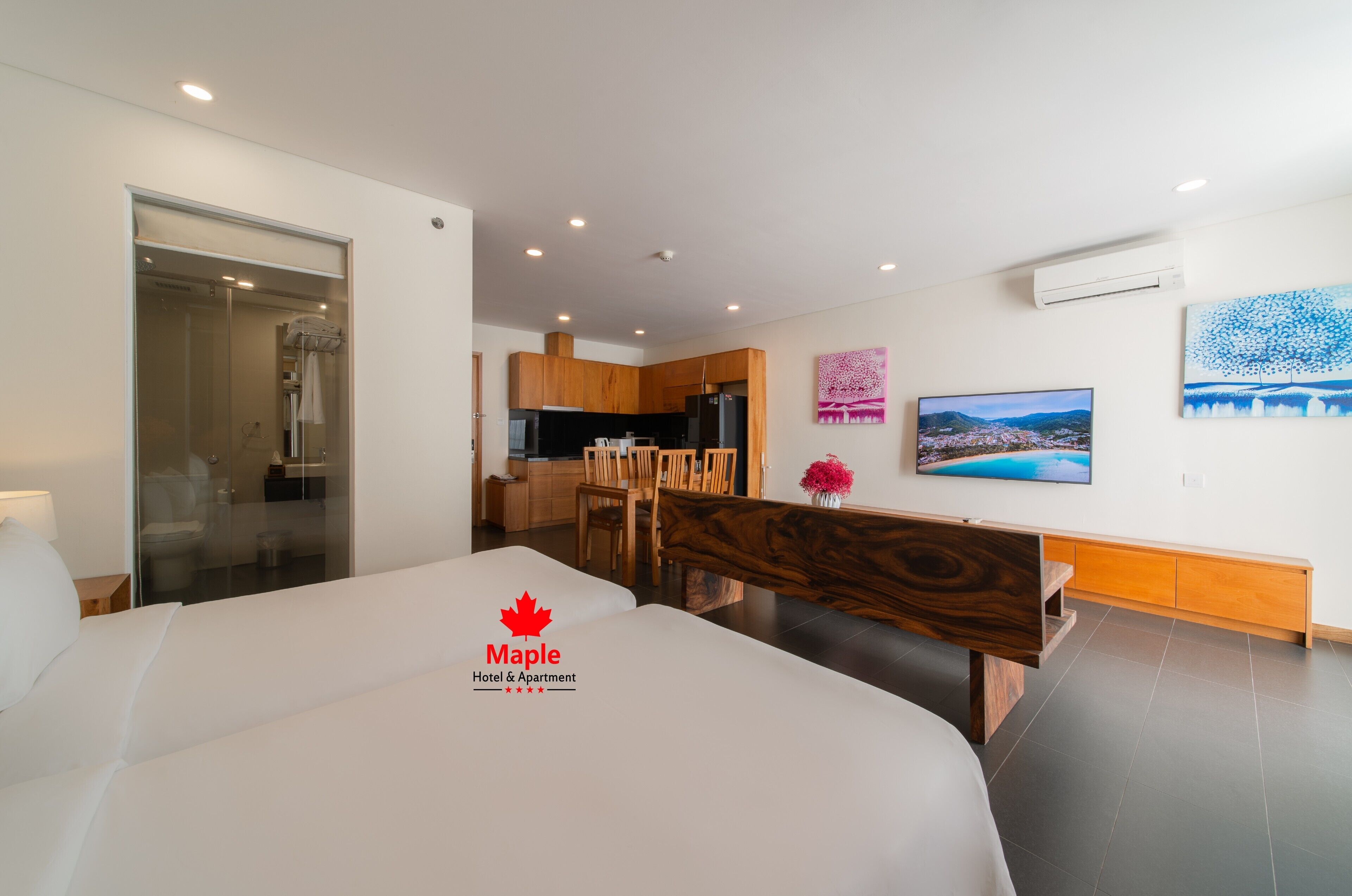 A modern hotel room with a large bed, a dining area with wooden furniture, and a kitchen in the background.