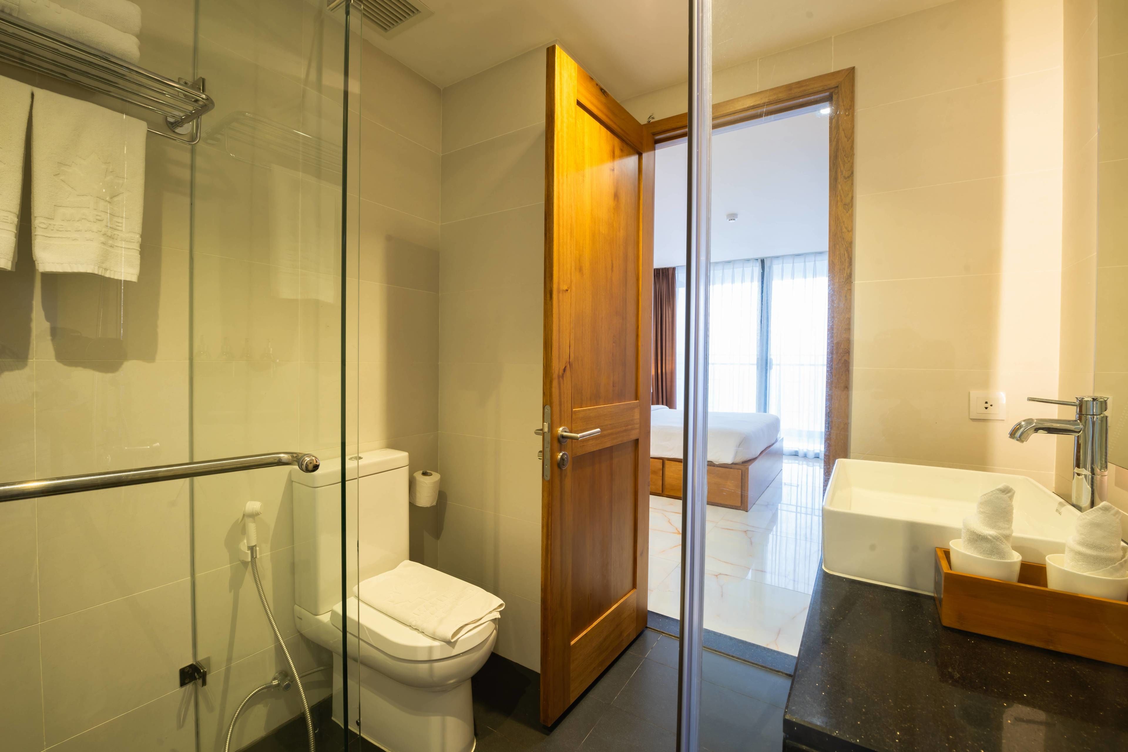 Een moderne badkamer met een douchecabine van glas, een wit toilet, een houten deur naar een slaapkamer en een badkuip met een houten dienblad.