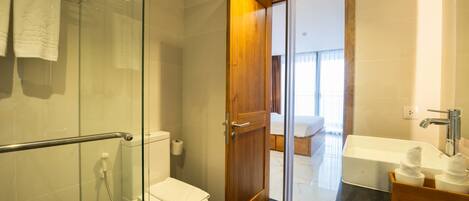 Een moderne badkamer met een douchecabine van glas, een wit toilet, een houten deur naar een slaapkamer en een badkuip met een houten dienblad.