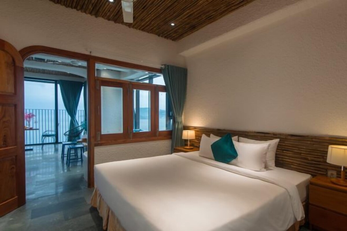Une chambre d’hôtel avec un grand lit, une tête de lit en bois et une vue sur un balcon extérieur, accessible par une porte vitrée.
