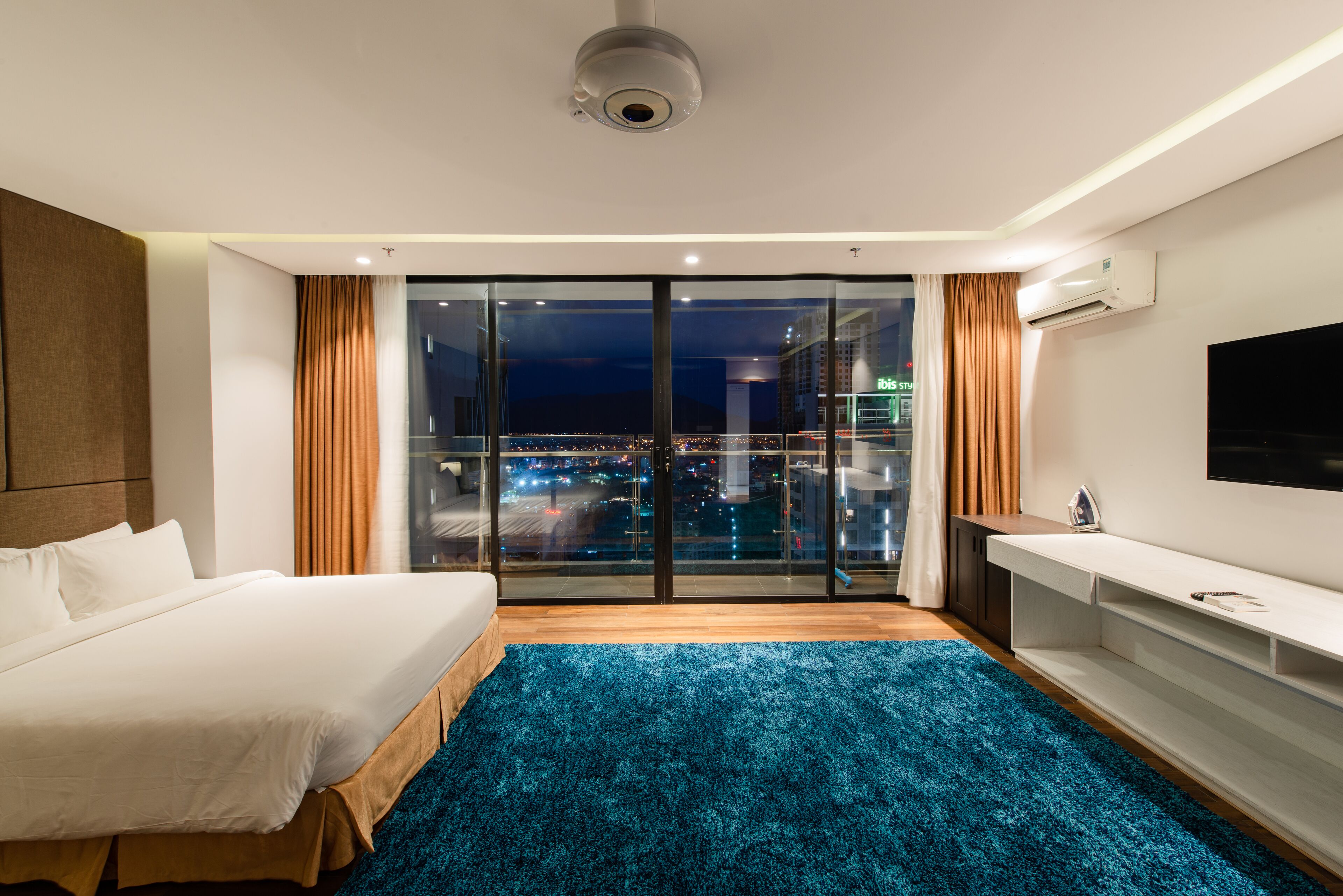 Una habitación de hotel moderna con una cama grande, una alfombra azul, un balcón con vistas a la ciudad y un televisor montado.