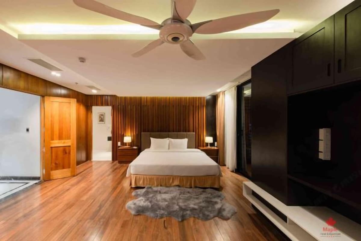 Une chambre moderne avec un plancher en bois, un lit recouvert de draps blancs, un ventilateur de plafond et un téléviseur posé sur un meuble.