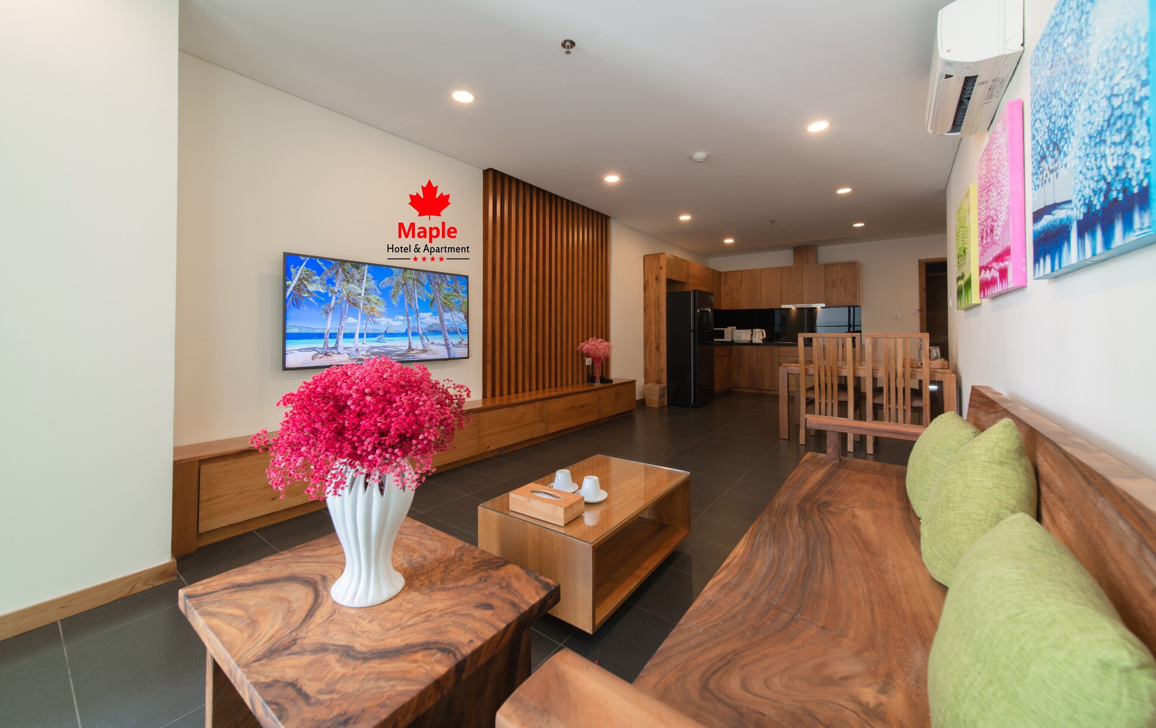 A modern hotel lobby with a wooden reception desk, a seating area with a sofa and chairs, a kitchen area in the background, and a large wall-mounted TV displaying a tropical beach scene.