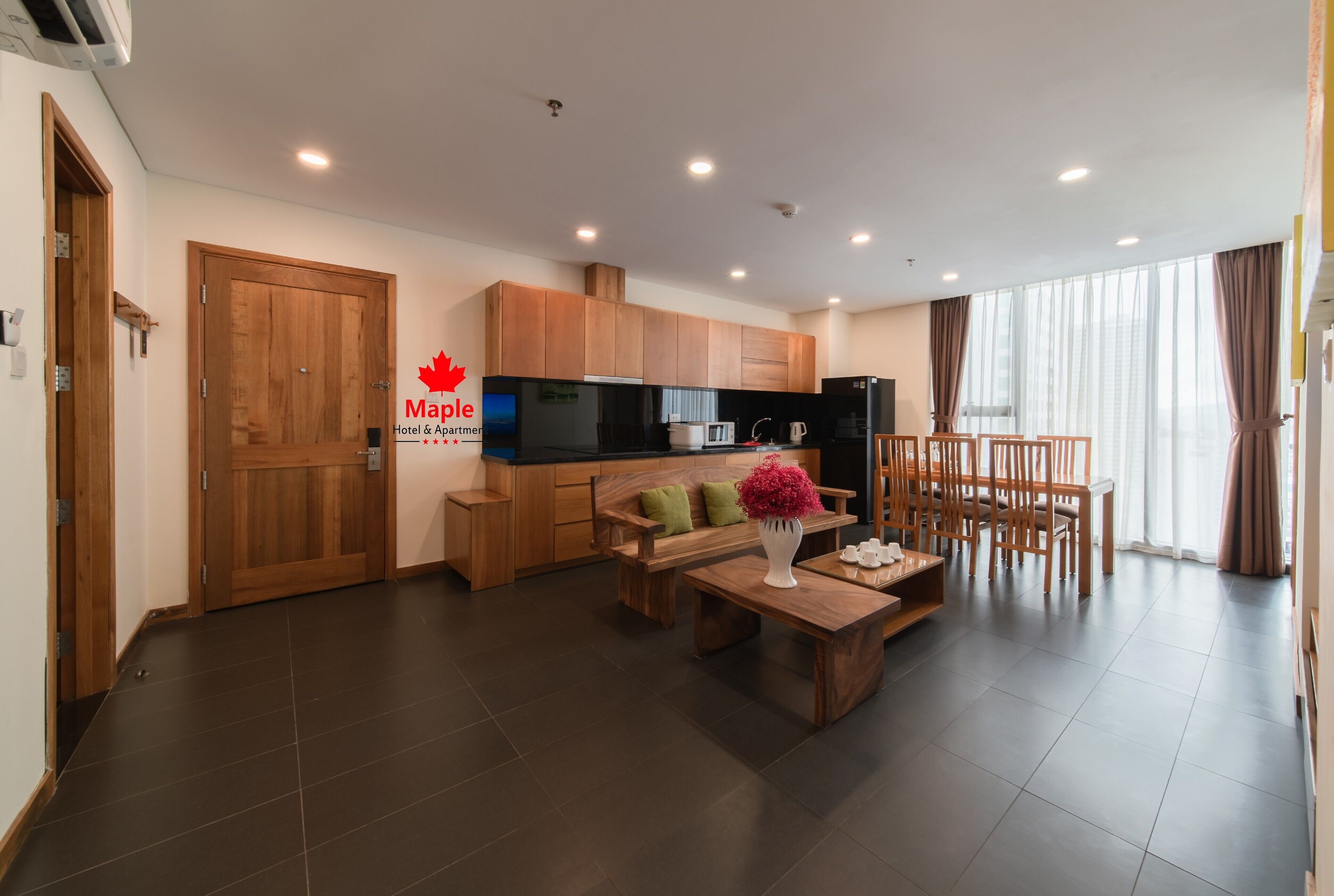 Une chambre d’hôtel moderne dotée d’une porte en bois, d’une kitchenette, d’un coin repas avec une table et des chaises, et d’une grande fenêtre avec des rideaux.